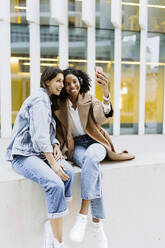 Happy young woman taking selfie with friends sitting on wall in front of building - JJF00628