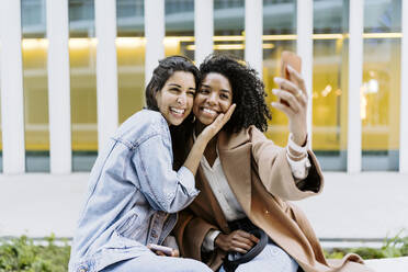 Happy friends taking selfie through smart phone in front of building - JJF00629