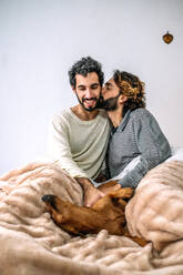 Schwuler Mann küsst Freund und spielt mit Hund auf dem Bett zu Hause - GDBF00046