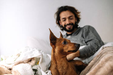 Smiling man playing with pet dog on bed at home - GDBF00047