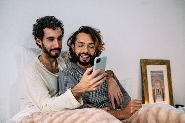 Mann teilt Smartphone mit Freund, der zu Hause auf dem Bett sitzt - GDBF00049