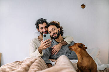 Lächelnde Homosexuell Paar beobachten Smartphone sitzen auf dem Bett zu Hause - GDBF00051