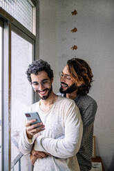 Happy gay man embracing boyfriend using smart phone near window at home - GDBF00052