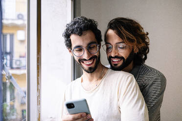 Man sharing smart phone with gay friend near window at home - GDBF00053