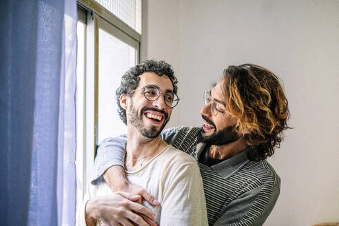 Happy gay man with arm around boyfriend near window at home - GDBF00054