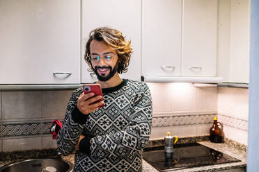 Smiling man using mobile phone in kitchen at home - GDBF00055