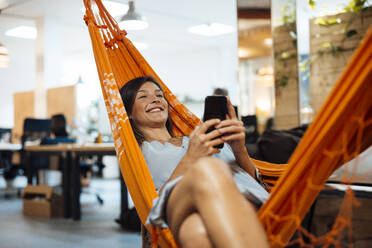 Lächelnde Geschäftsfrau mit Smartphone entspannt in Hängematte im Büro - JOSEF17564