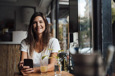 Happy woman sitting with smart phone in cafe - JOSEF17598