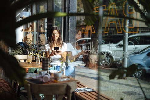 Frau, die durch ein Glasfenster in einem Café eine Textnachricht über ihr Smartphone sendet - JOSEF17600