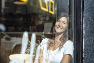Lächelnde Frau, die sich in einem Café an ein Fenster lehnt, gesehen durch Glas - JOSEF17602
