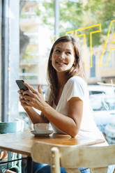 Smiling woman holding smart phone sitting with coffee cup at table in cafe - JOSEF17616