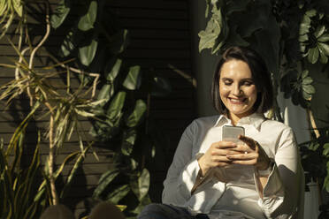 Glückliche Frau, die auf dem Balkon mit ihrem Mobiltelefon im Internet surft - NJAF00281