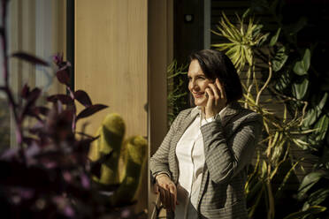 Lächelnde Geschäftsfrau, die auf einem sonnigen Balkon mit Pflanzen mit ihrem Smartphone telefoniert - NJAF00284