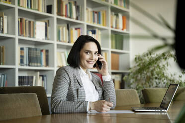 Smiling businesswoman talking on smart phone sitting at desk in office - NJAF00285