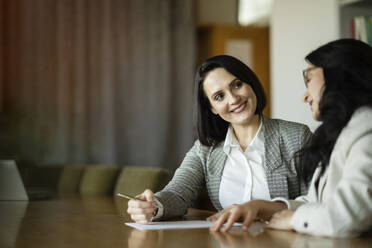 Smiling colleague looking at businesswoman at desk in office - NJAF00295