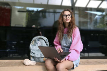 Contemplative student with laptop and backpack sitting on bench - SYEF00284