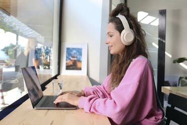 Geschäftsfrau mit drahtlosen Kopfhörern bei der Arbeit am Laptop in einem Café - SYEF00286