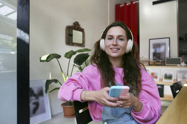 Lächelnder Freiberufler mit drahtlosen Kopfhörern und Smartphone in einem Café - SYEF00302