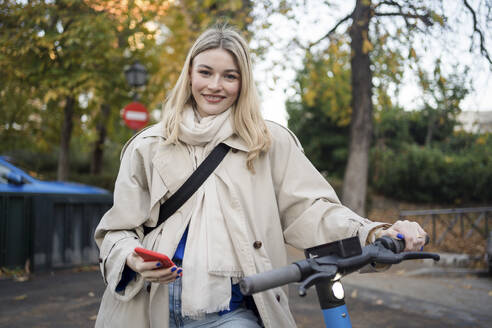 Lächelnde Frau mit Elektroroller, die ein Mobiltelefon hält - JJF00654