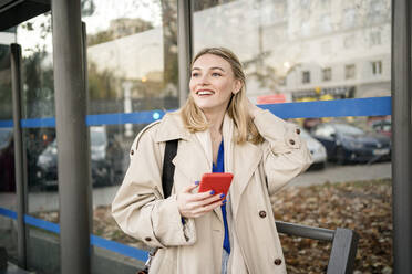 Glückliche junge Frau mit Hand im Haar, die an der Bushaltestelle ihr Smartphone hält - JJF00670