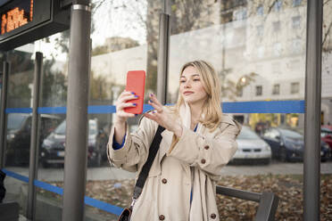 Eine junge Frau fängt einen Moment der Selbstdarstellung und der Verbundenheit mit ihrer Umgebung durch ein Smartphone-Selfie an einer Bushaltestelle ein - JJF00671