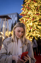 Young woman using smart phone in front of illuminated Christmas tree at night - JJF00678