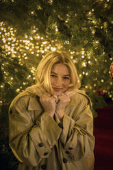 Lächelnde blonde Frau unter beleuchtetem Weihnachtsbaum bei Nacht - JJF00687