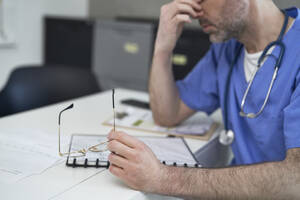 Müder reifer Arzt sitzt am Schreibtisch in der Klinik - ABIF01909