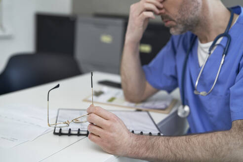 Tired mature doctor sitting at desk in clinic - ABIF01909