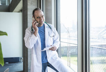 Lächelnder reifer Arzt, der am Fenster steht und mit einem Smartphone spricht - UUF28475