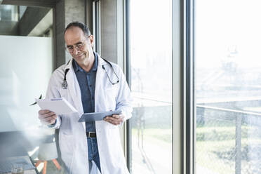 Smiling mature doctor reading prescription and holding tablet PC by window - UUF28482