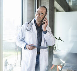 Älterer Arzt mit Tablet-PC im Gespräch mit Smartphone am Fenster - UUF28483