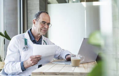 Ein Arzt hält einen medizinischen Bericht und benutzt einen Laptop am Schreibtisch - UUF28484