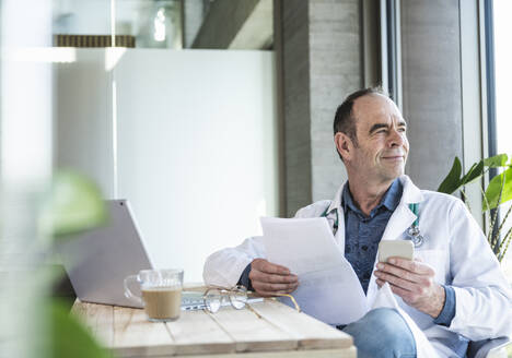 Nachdenklich lächelnder Arzt sitzt mit medizinischem Bericht und Smartphone am Schreibtisch - UUF28485