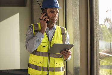 Engineer using walkie talkie standing with tablet PC in office - UUF28519