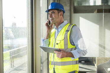 Thoughtful engineer talking on mobile phone looking through window in office - UUF28526