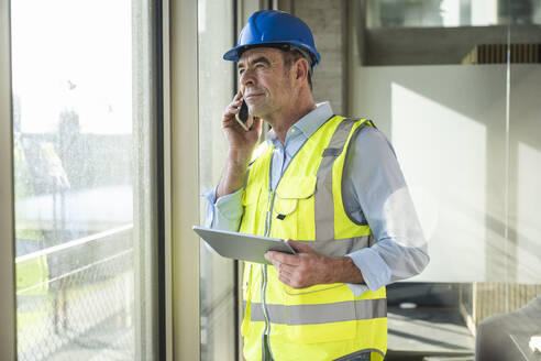 Nachdenklicher Ingenieur, der mit seinem Handy telefoniert und durch ein Fenster im Büro schaut - UUF28526