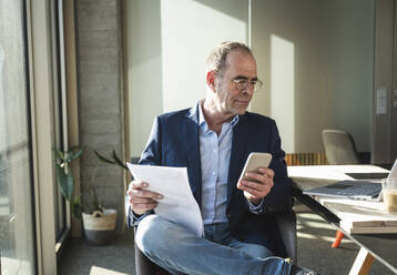 Älterer Geschäftsmann, der ein Smartphone benutzt und mit einem Dokument im Büro sitzt - UUF28534