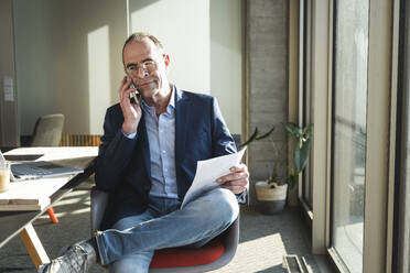 Älterer Geschäftsmann, der mit einem Smartphone spricht und ein Dokument im Büro hält - UUF28535