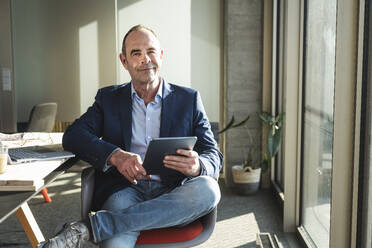 Confident mature businessman sitting with tablet PC in office - UUF28536