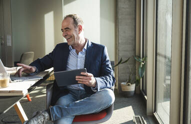 Happy businessman using laptop and holding tablet PC in office - UUF28537