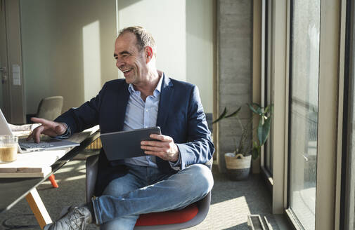 Glücklicher Geschäftsmann, der einen Laptop benutzt und einen Tablet-PC im Büro hält - UUF28537