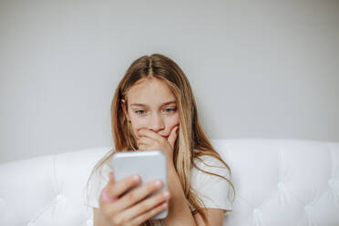 Worried girl using smart phone sitting on bed at home - MDOF00863