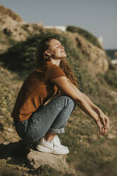 Woman with eyes closed crouching on rock at sunny day - DMGF01089