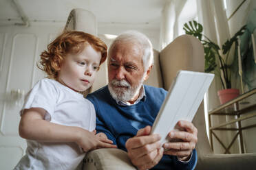 Großvater teilt Tablet-PC mit Enkel, der zu Hause im Sessel sitzt - MDOF00877