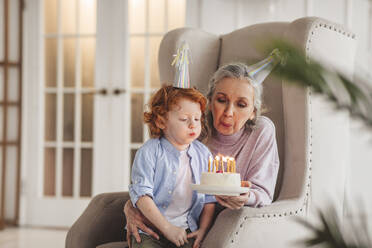 Enkel und Großmutter tragen einen Partyhut und blasen Kerzen auf einer Geburtstagstorte zu Hause aus - MDOF00881