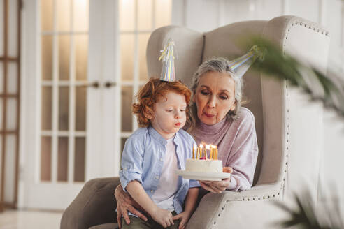 Grandson and grandmother wearing party hat and blowing candles on birthday cake at home - MDOF00881