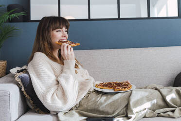 Happy woman enjoying eating pizza sitting on sofa at home - PNAF05046