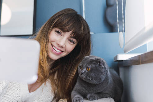 Freudige Frau fängt einen schnurrigen Moment mit ihrer Katze ein, während sie zu Hause ein Selfie schießt - PNAF05054