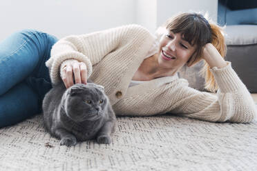 Lächelnde Frau streichelt Katze, die zu Hause auf dem Teppich liegt - PNAF05055